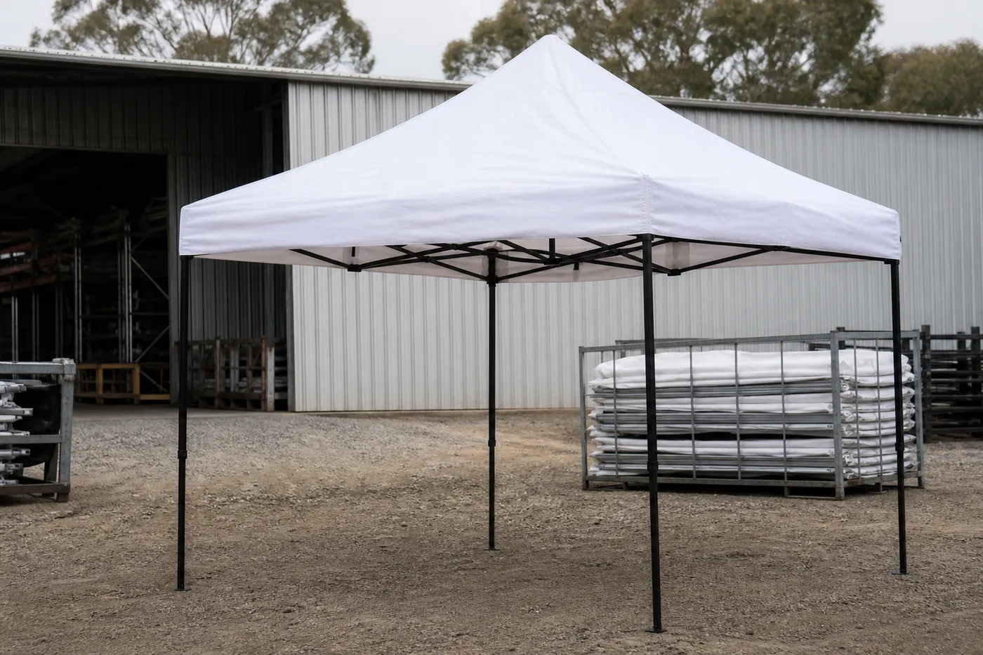 White square pop-up canopy on a gravel yard in front of a corrugated shed, with folded roof fabric stacked in a metal crate.