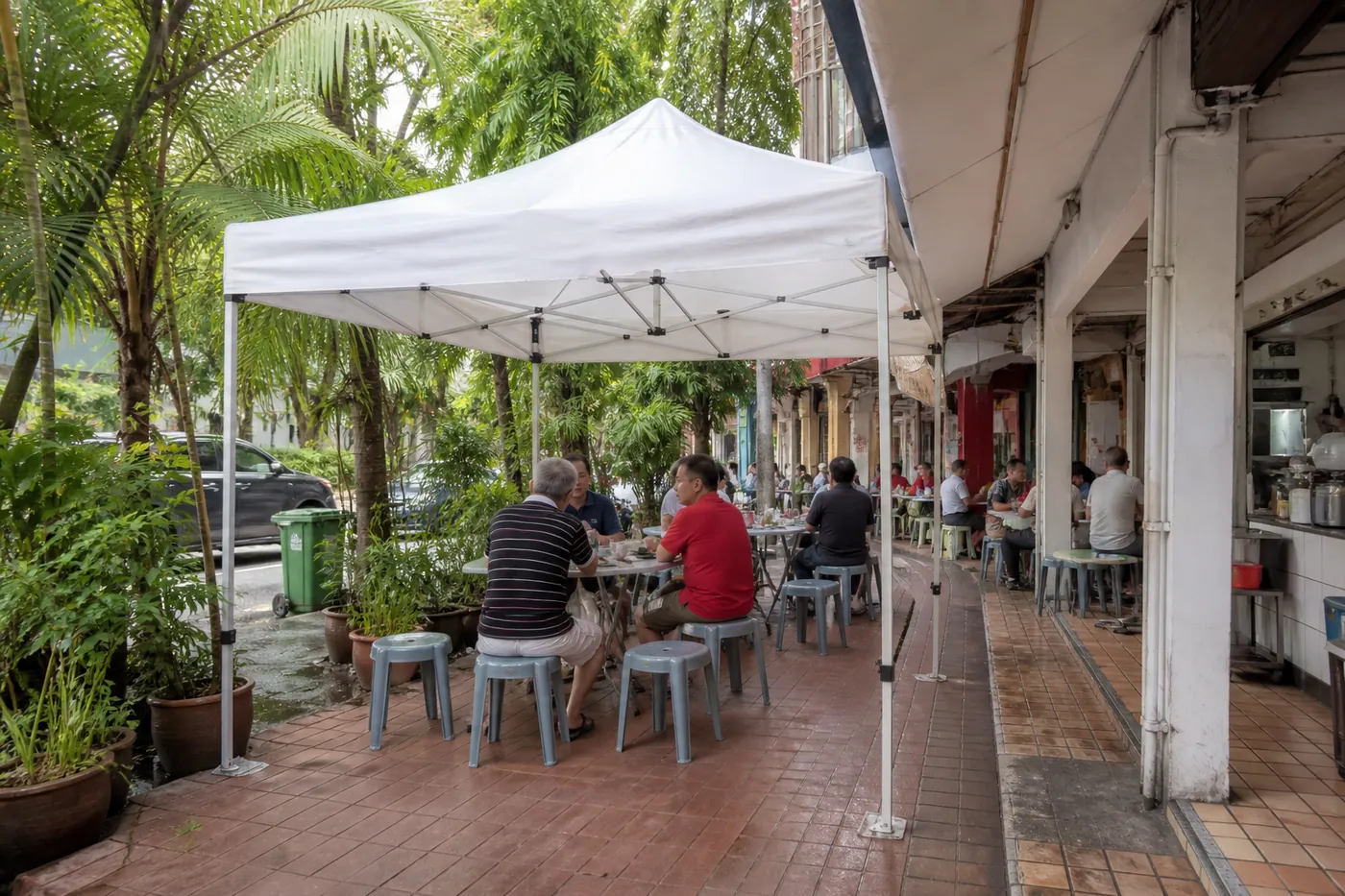 White pop-up canopy on a red-brick terrace beside a shophouse walkway, with round tables, stools and tropical planters.