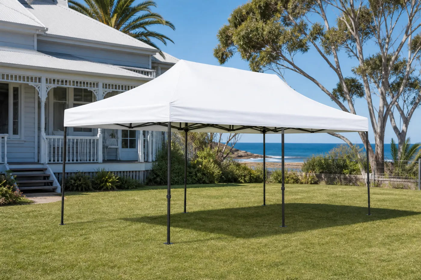 Rectangular white pop-up canopy on a lawn in front of a coastal weatherboard house, with the ocean beyond the trees.
