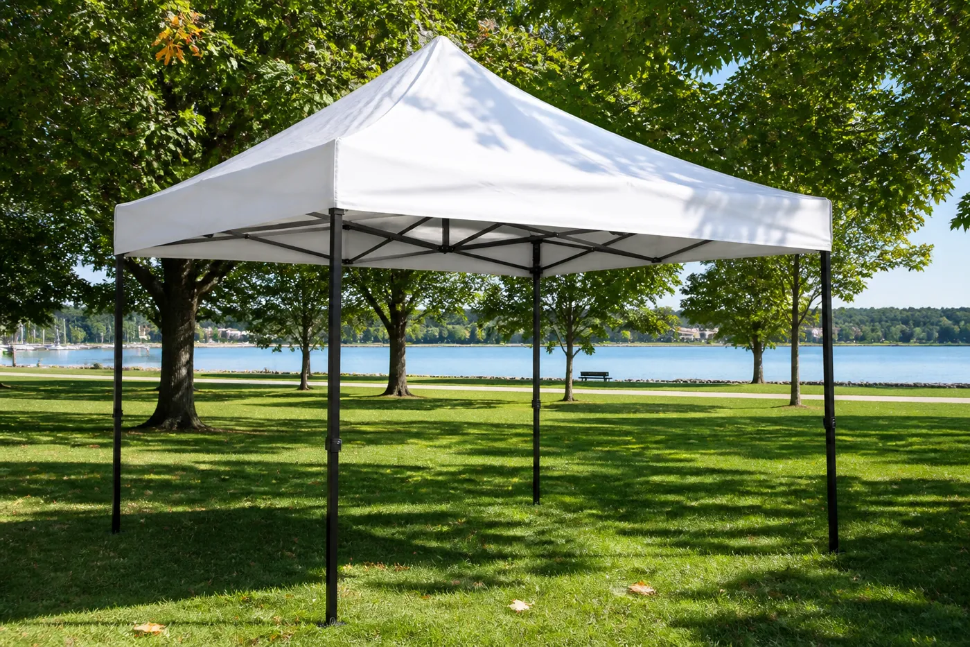 White square pop-up canopy on a lakeside park lawn, with deciduous trees and water behind it.