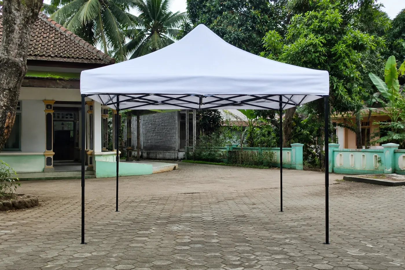 White square pop-up canopy on hexagonal courtyard pavers, with tropical plants and a tiled-roof house behind it.