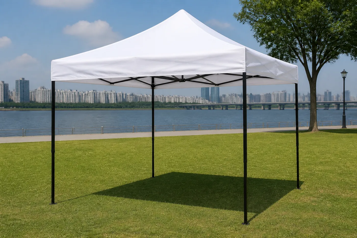 White square pop-up canopy on a riverside park lawn, with a city skyline and bridge across the water.