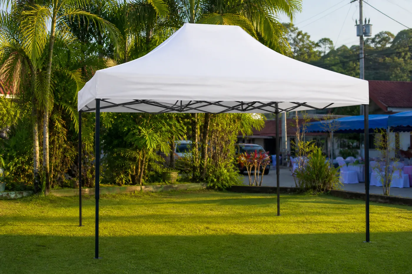 White square pop-up canopy on a tropical garden lawn, with palms and a larger blue event tent in the background.