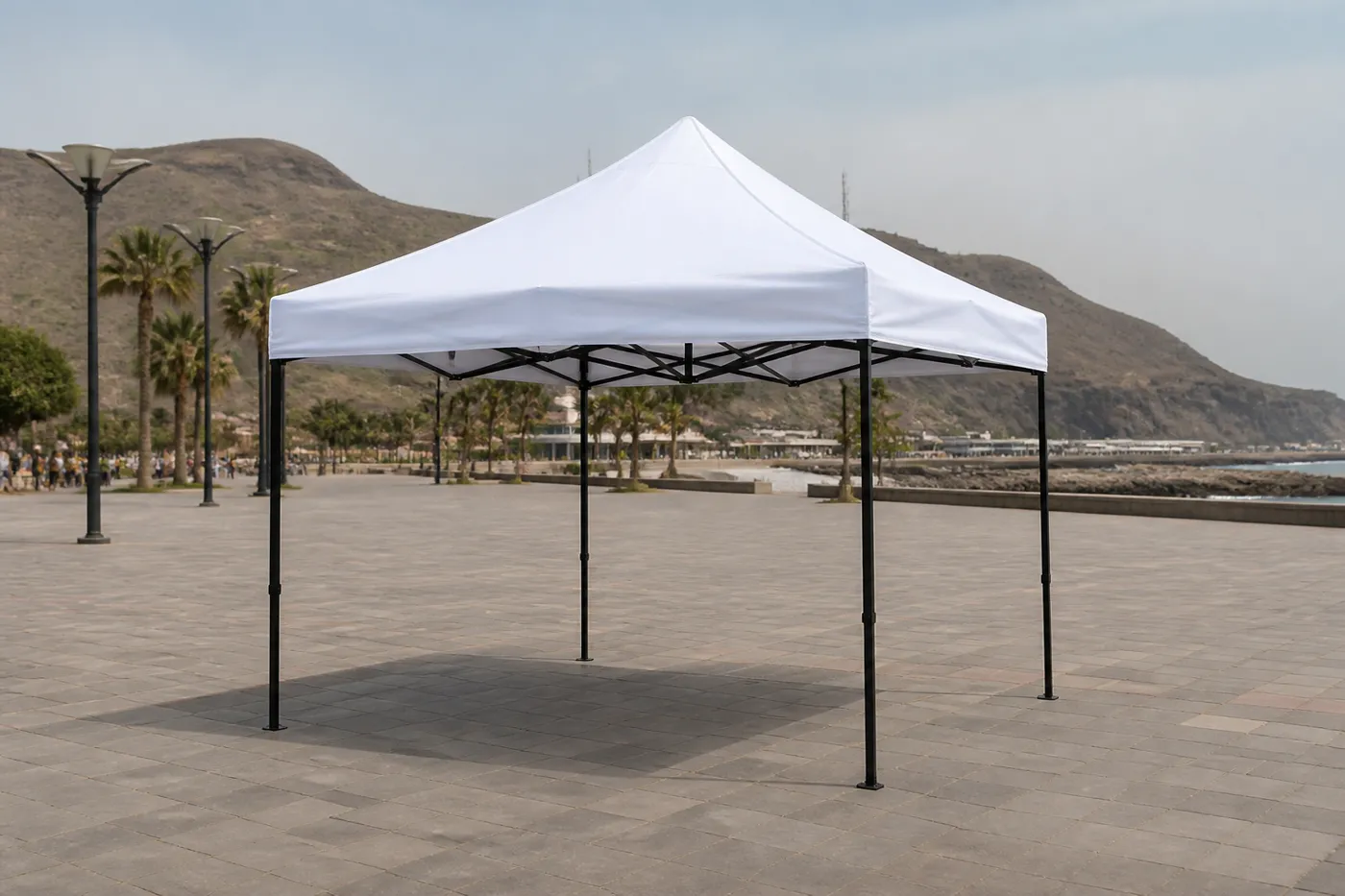 White square pop-up canopy on a paved coastal promenade, with palms, streetlights and a dry mountain by the sea.
