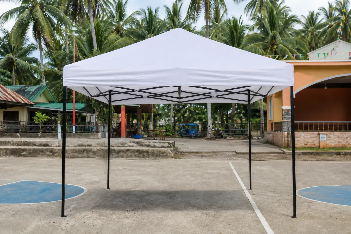White square pop-up canopy on a barangay basketball court, with coconut palms, a metal-roof house and pennant banners.