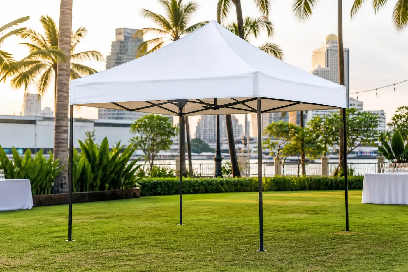 White rectangular pop-up canopy on a riverside lawn with palms and banquet tables, and a city skyline across the water.
