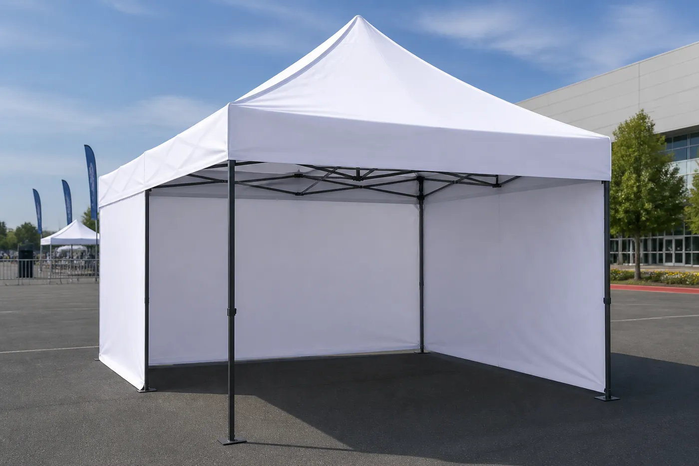 White pop-up canopy with side walls on an asphalt lot, with a modern building and distant event flags.