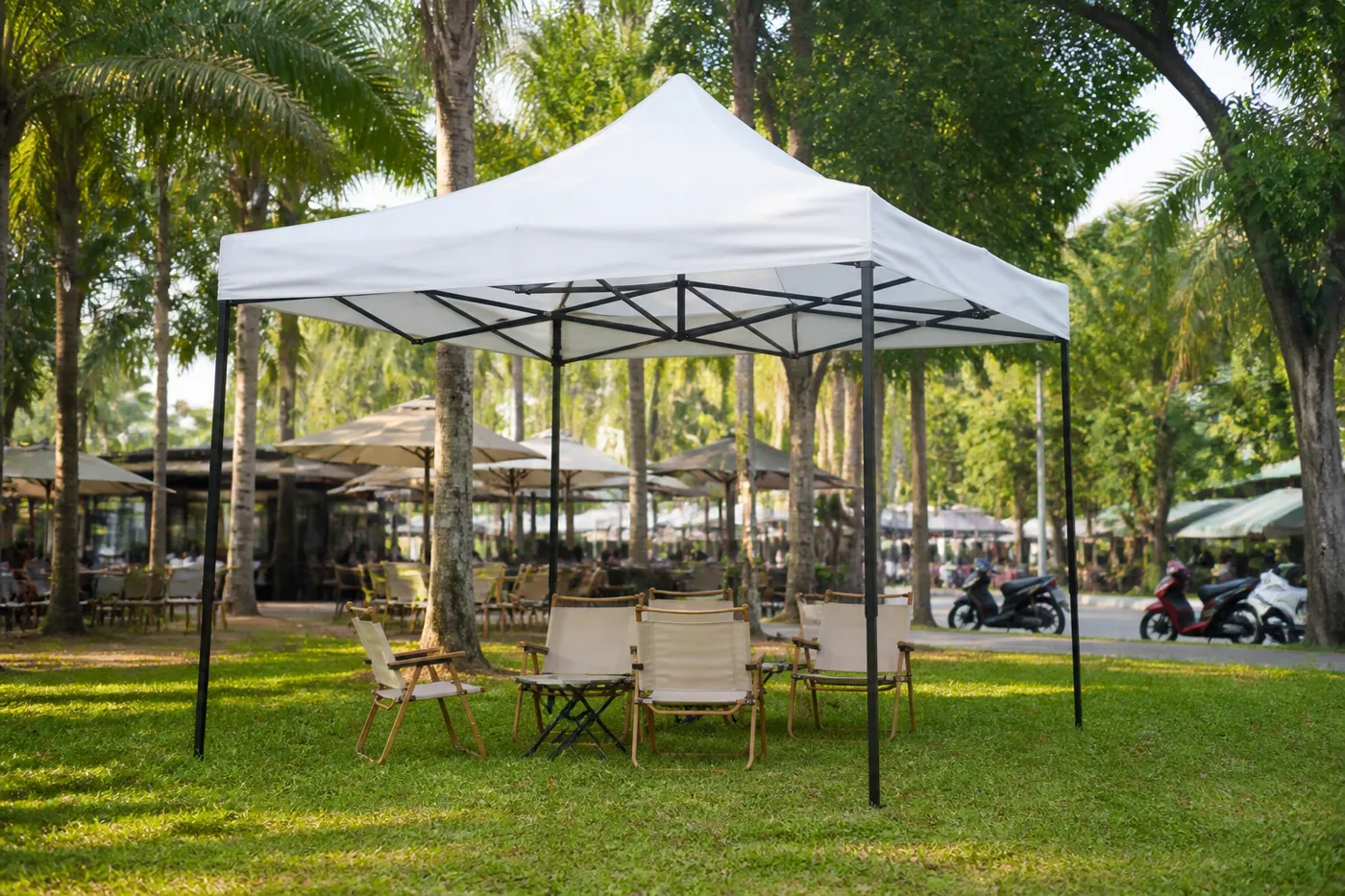 White square pop-up canopy on a tropical lawn, with folding chairs underneath and palms and parked motorcycles behind.